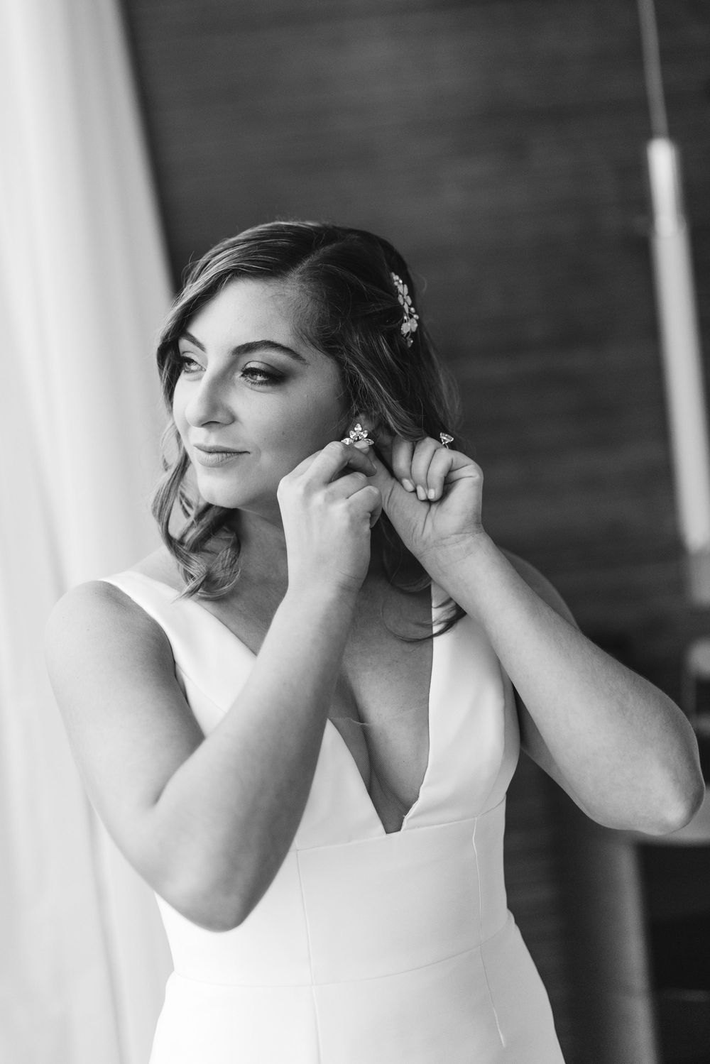Black and white portrait of a bride putting on earrings while wearing an elegant sleeveless wedding dress.