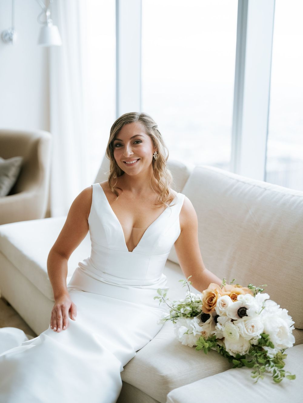 A bride in a white wedding dress sits on a couch holding a beautiful bouquet of white flowers.