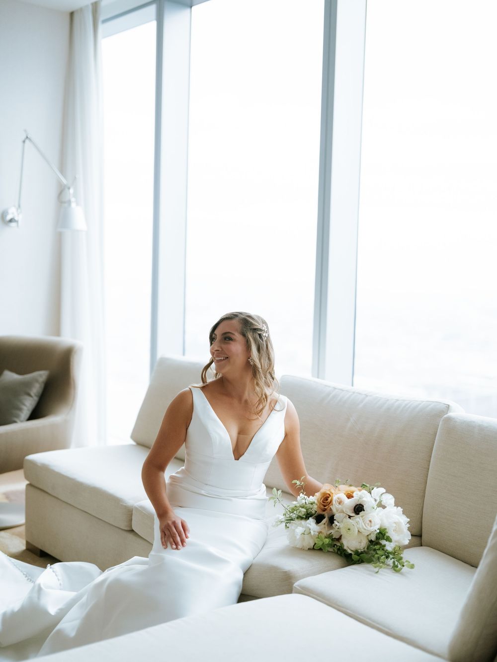 Elegant bride in white v-neck wedding dress sits on cream couch with floral bouquet near bright window.