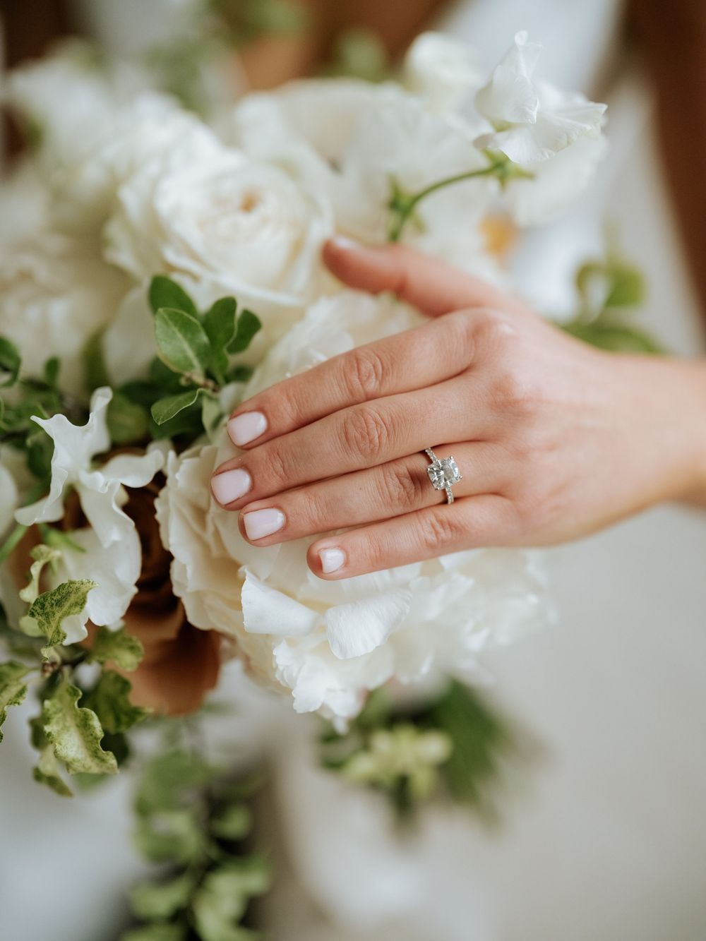 Close up of manicured hand with engagement ring touching white roses and greenery in bouquet.