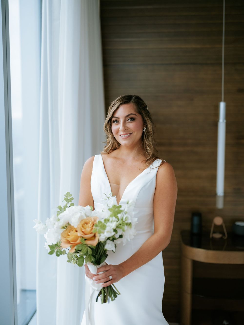 Bride in elegant white wedding dress holds a beautiful bouquet of peach and white flowers against wooden wall backdrop.