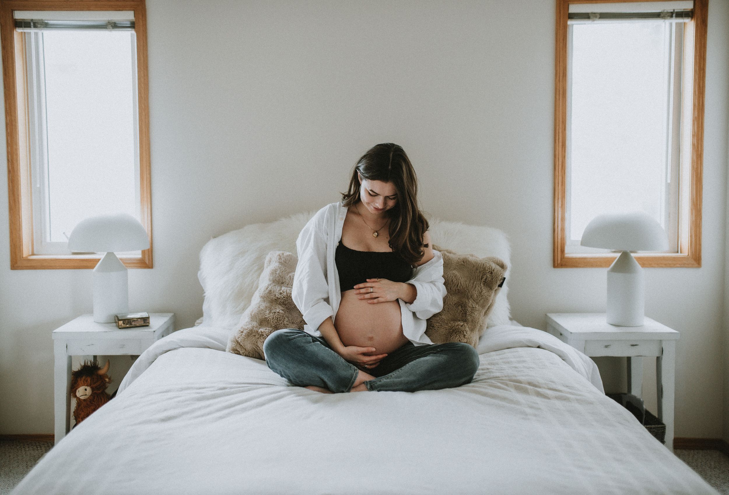 A mother to be cradles her pregnant belly as she sits on her bed in a bright bedroom during her maternity session in Cochrane Alberta