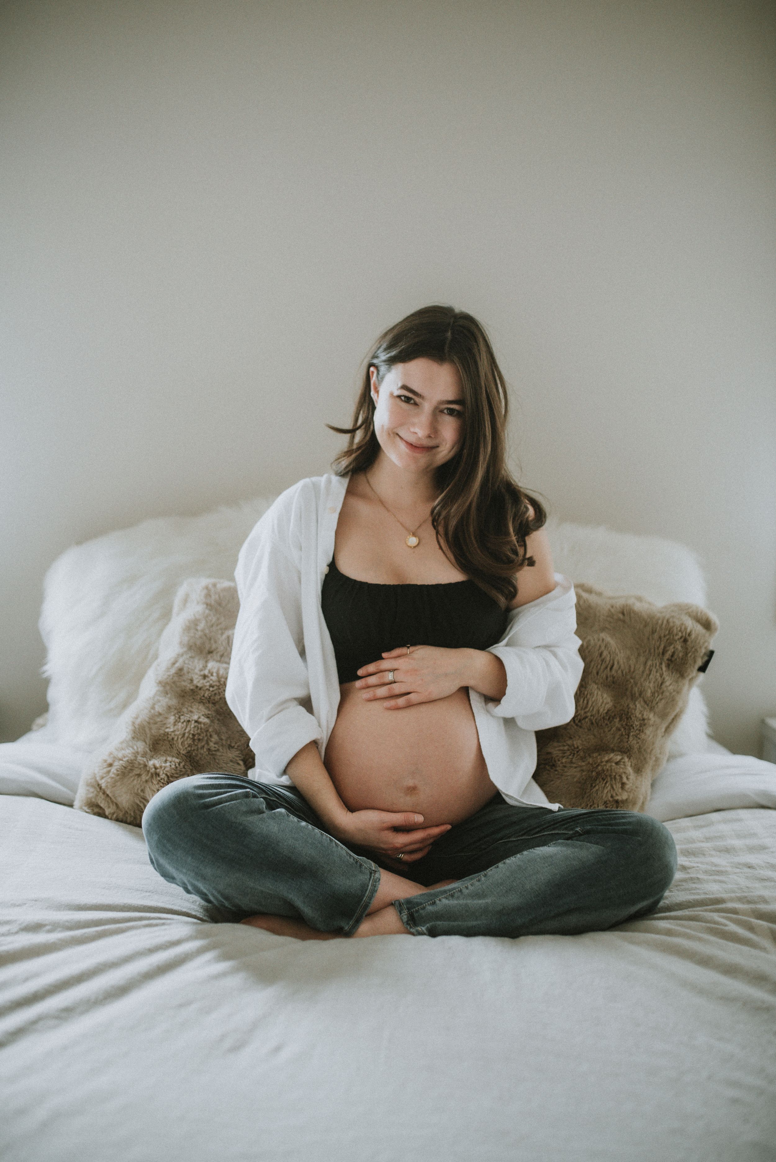 A mother to be cradles her pregnant belly as she sits on her bed in a bright bedroom during her maternity session in Cochrane Alberta