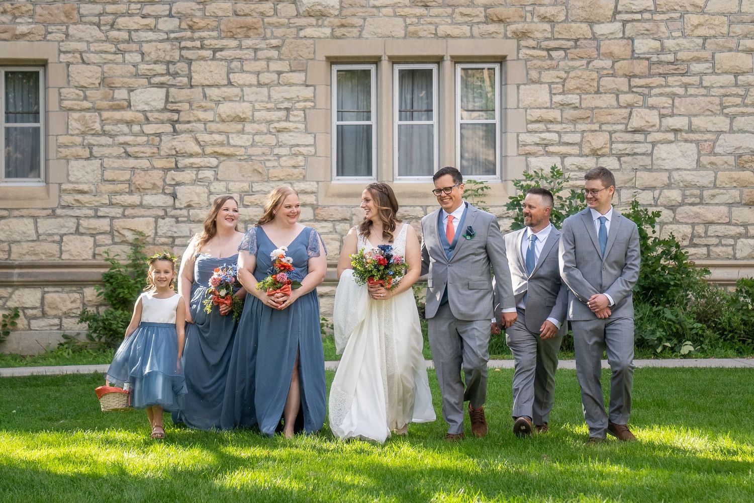 Wedding party members stand in a row against stone wall wearing gray and blue attire.