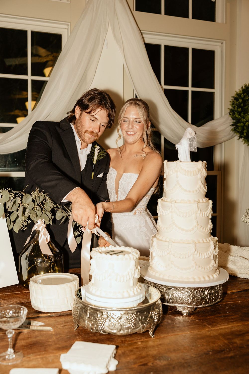 Newlyweds cut their elegant white tiered wedding cake together at indoor reception venue at The Elms of Coosada in Coosada, Alabama