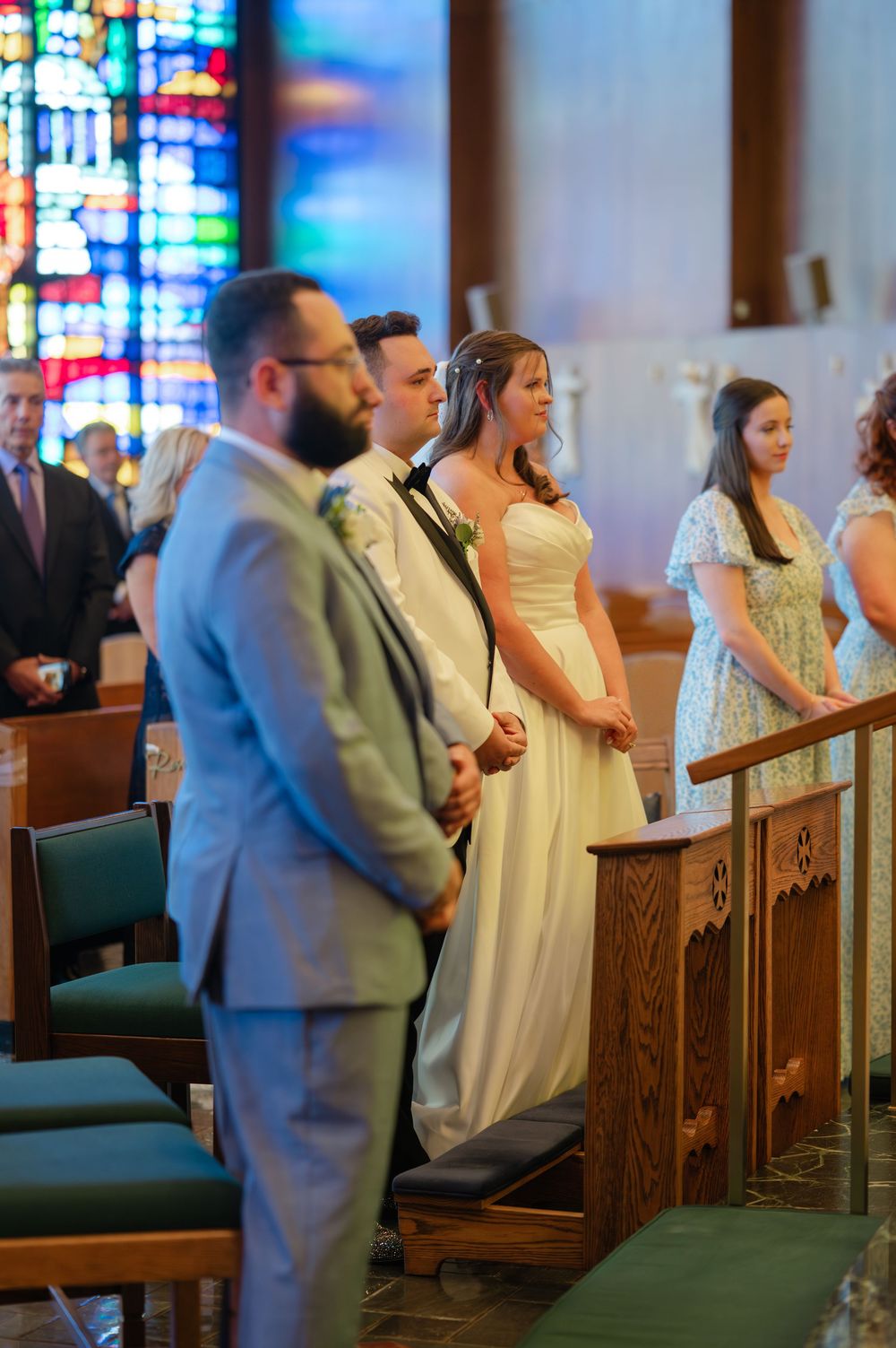 Wedding ceremony in a church with stained glass windows and guests standing in formal attire.