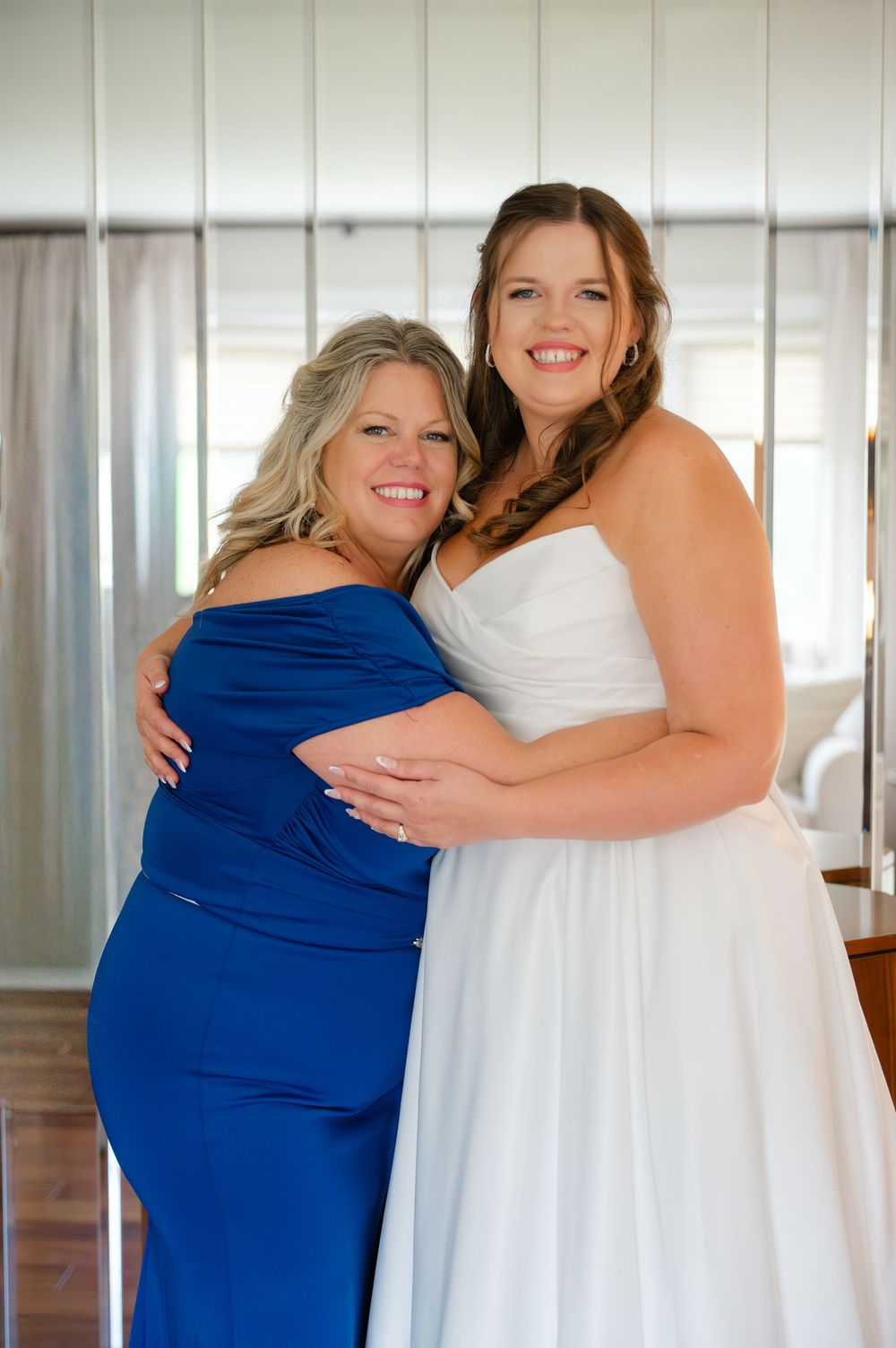 Two women share an emotional embrace, one wearing a royal blue dress and the other in a white wedding gown.