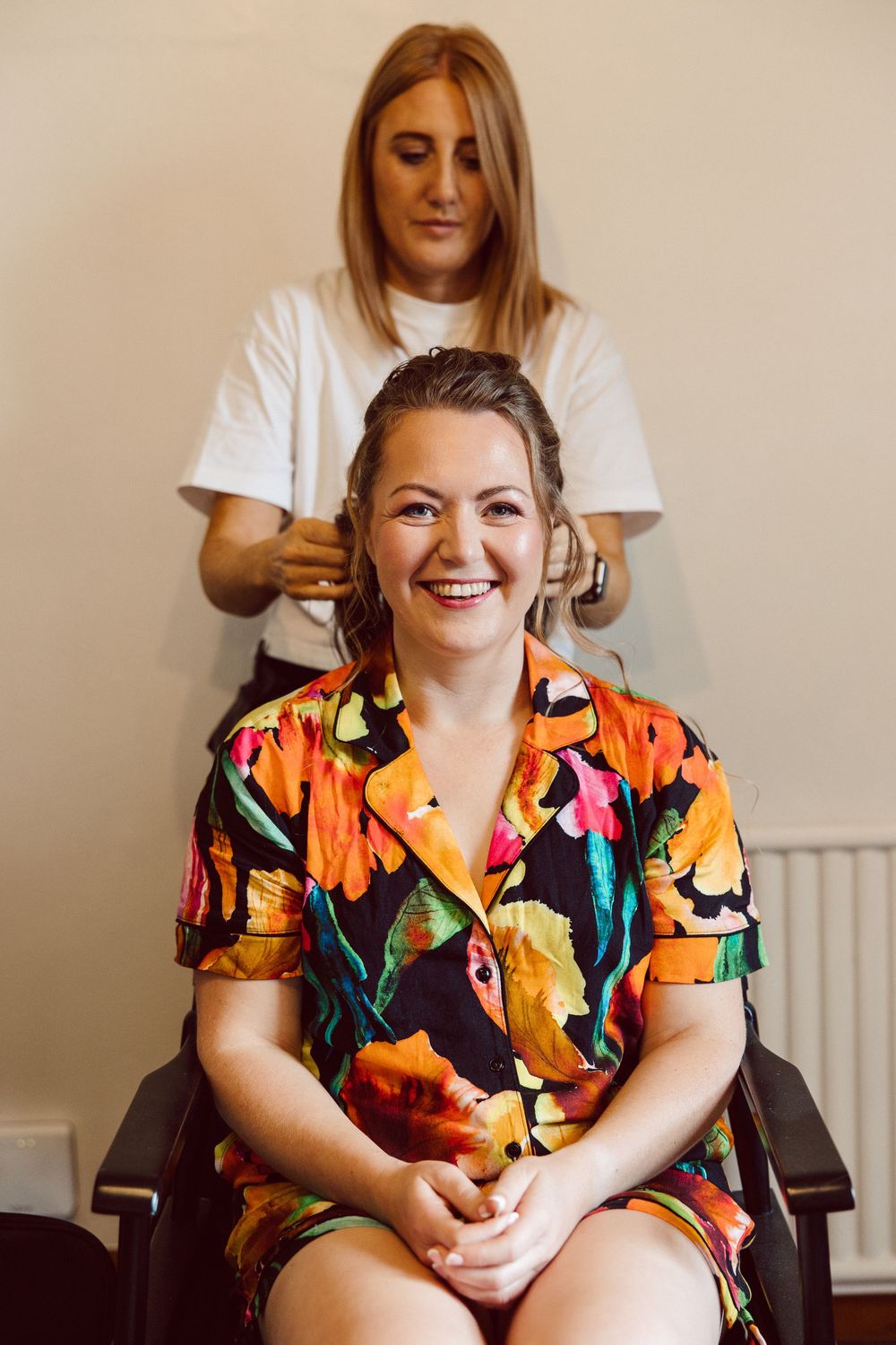 A person in a vibrant floral print shirt sits smiling while getting their hair styled in a bright indoor setting.