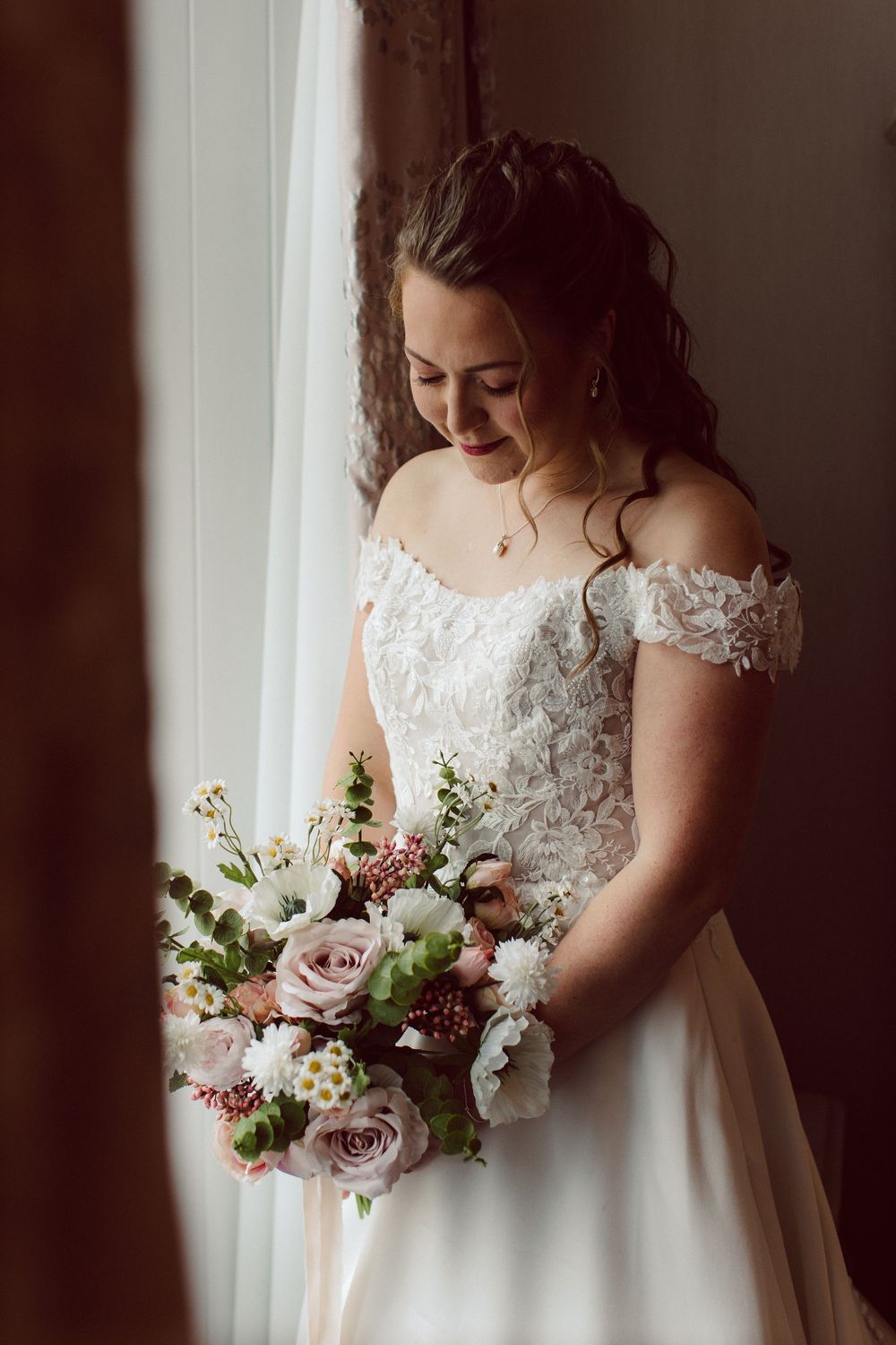 Beautiful bridal portrait showing an off-shoulder lace wedding dress and romantic pink and white floral bouquet by a window.