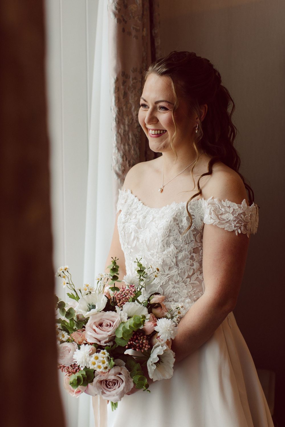 A bride in a white lace wedding dress holds a romantic bouquet of pink roses and white flowers near a window.