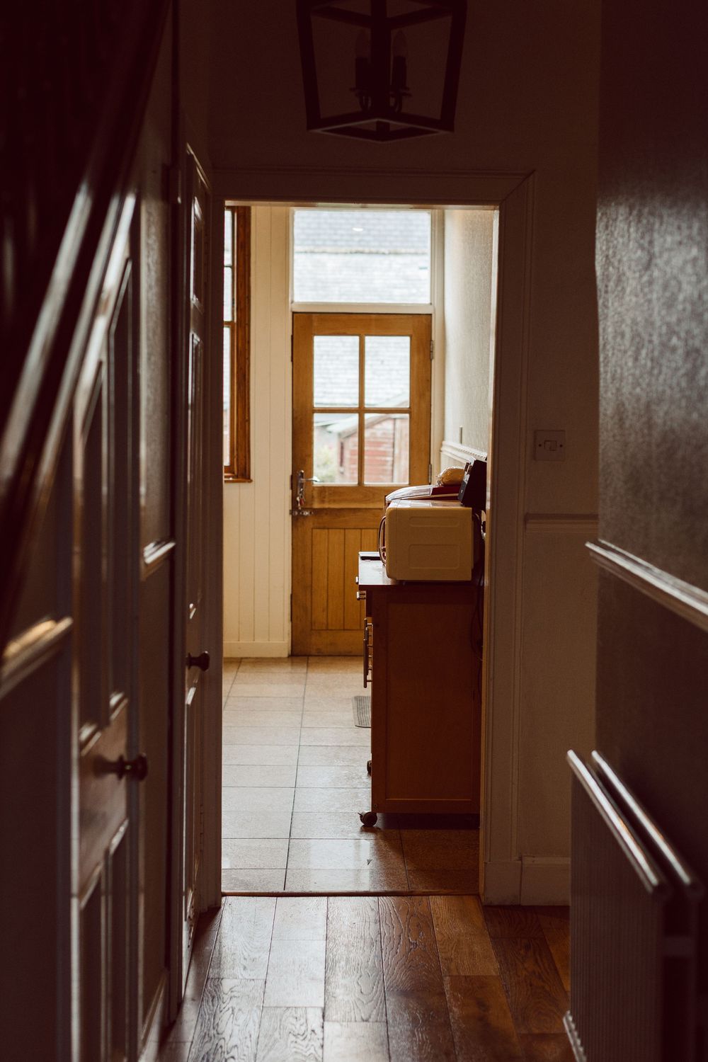 Sunlight streams through windows into an empty hallway with tiled floors and wooden doors creating dramatic shadows.