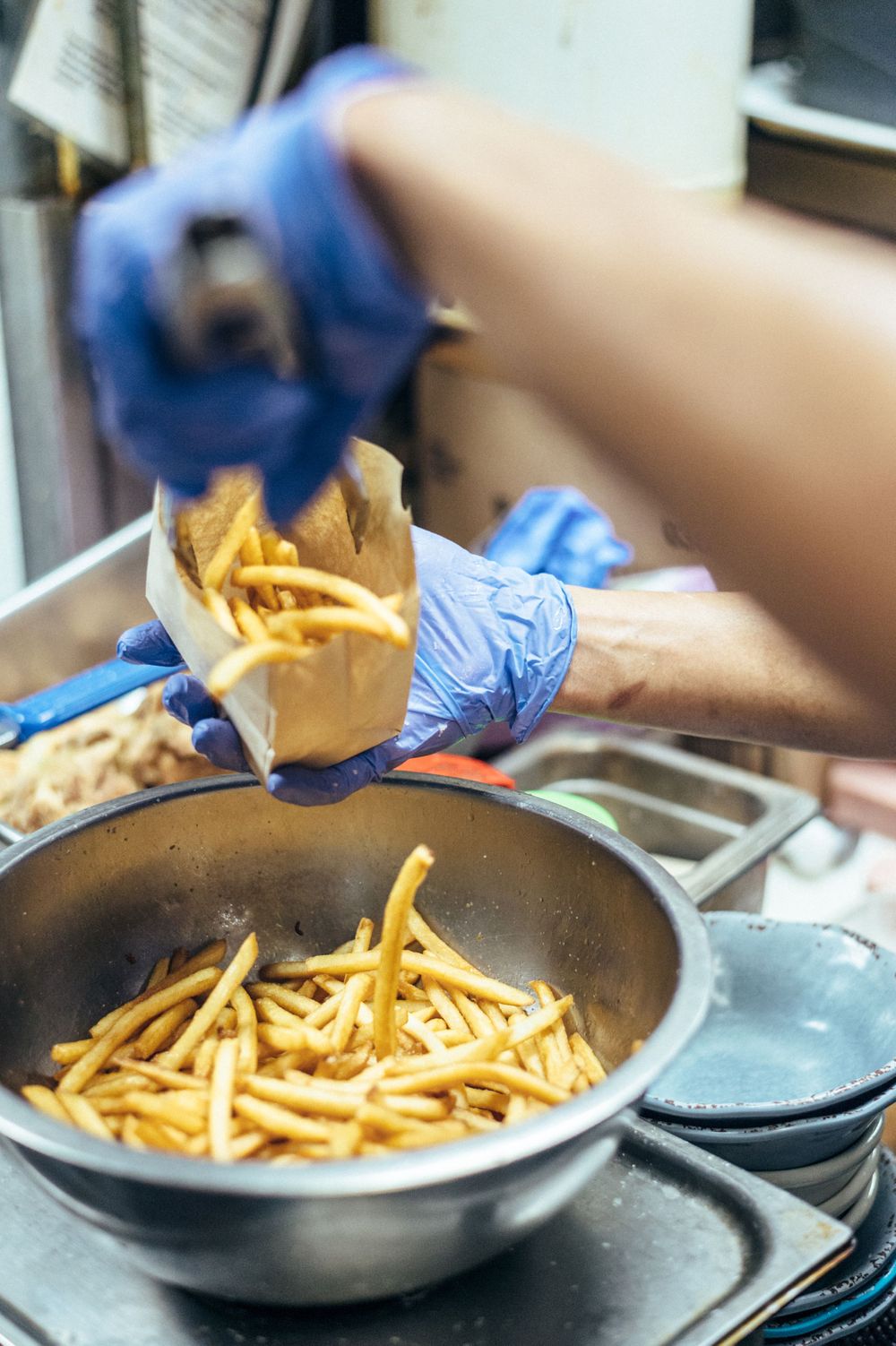 Chef tossing french fries in a stainless steel pan while wearing blue gloves.