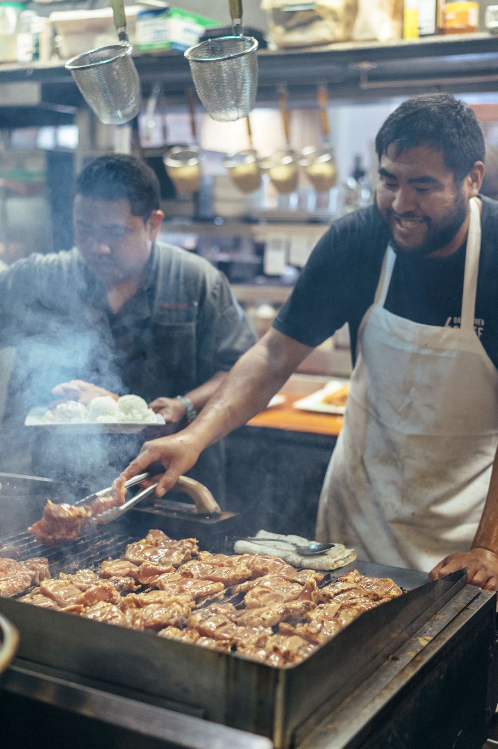 Cooks grilling meat on a large commercial flat top grill with smoke rising.