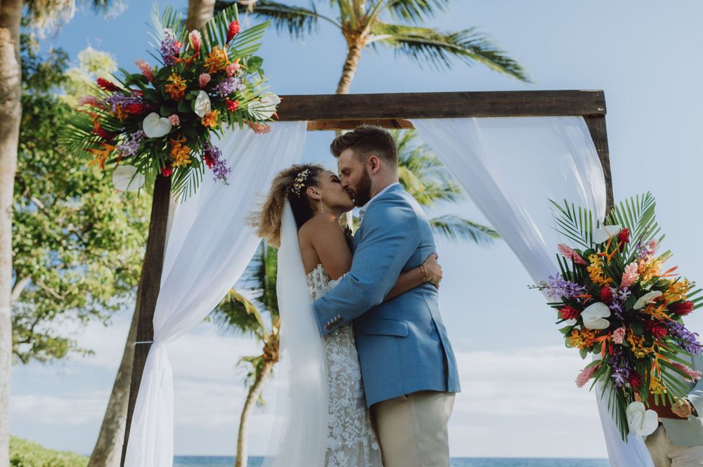 Couple shares first kiss under decorated arch at beachside wedding ceremony.