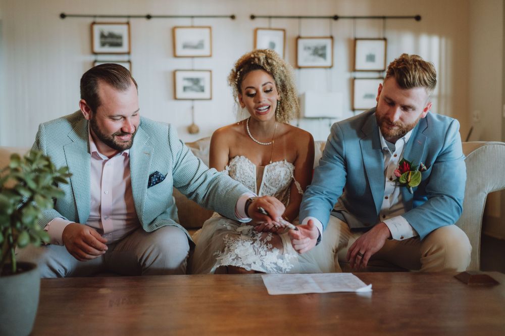Couple signing marriage documents at a wooden table indoors with framed artwork on wall.