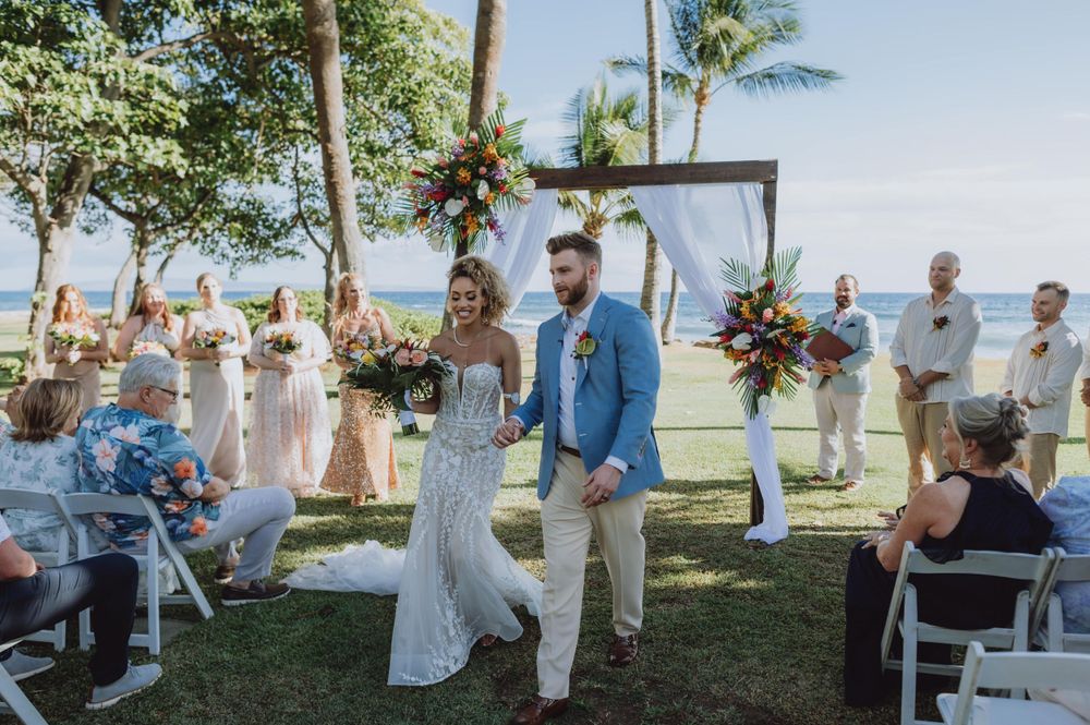 Wedding guests observe ceremony from white chairs at tropical outdoor venue.