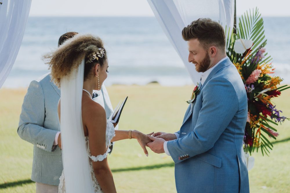 Couple exchanges vows during a beachside ceremony under a white fabric arch.