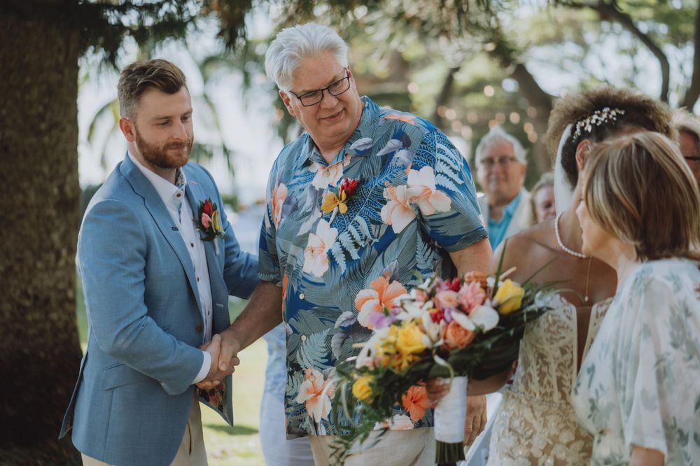 Wedding ceremony participants share emotional moments under tropical trees.