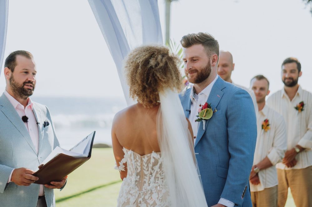 Wedding ceremony takes place under a white arch with ocean views and tropical decor.