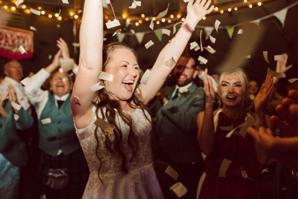 Wedding guests dancing and celebrating with arms raised on a festive dance floor illuminated by string lights.