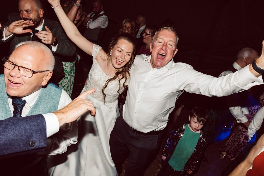 Energetic crowd dancing and laughing at a wedding reception in a dark venue with ambient lighting.
