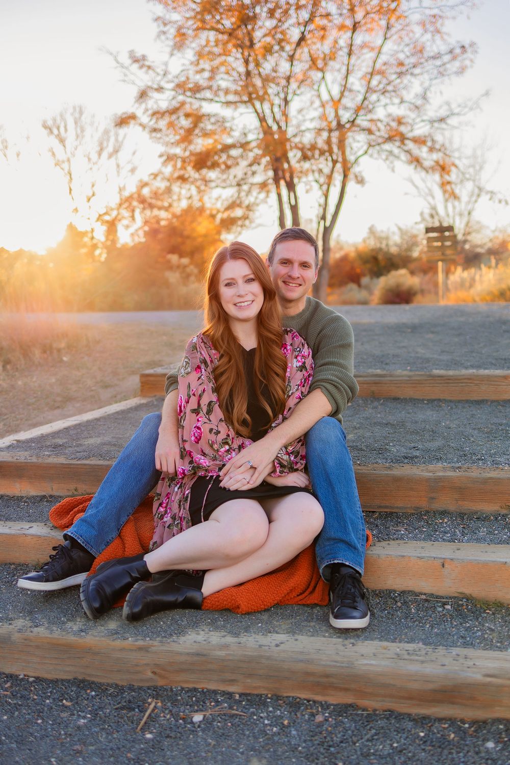 A couple sits together on gravel stairs at sunset while wearing casual clothing and embracing.