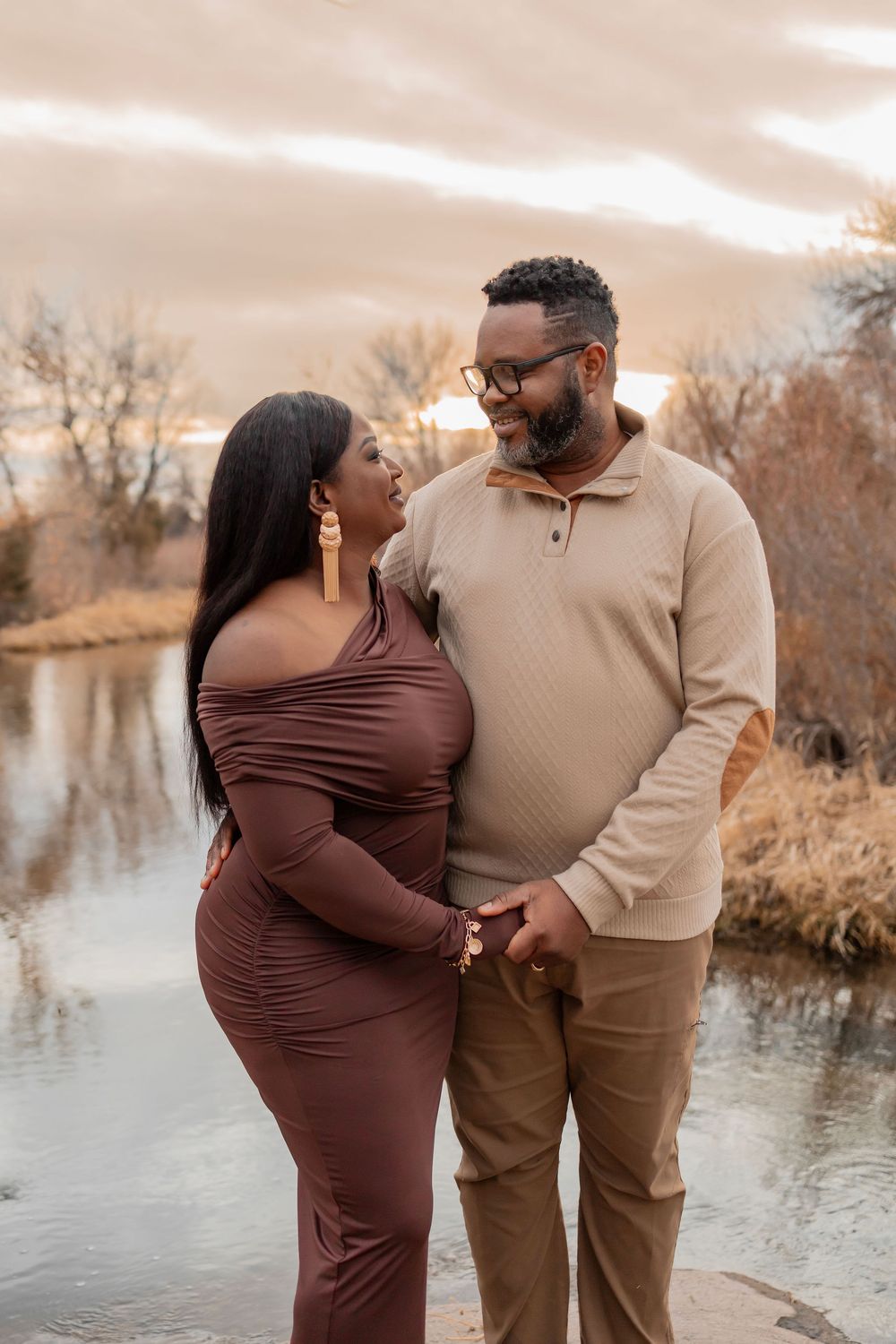 A couple in brown and beige outfits share intimate moments by a serene lake during sunset in autumn.