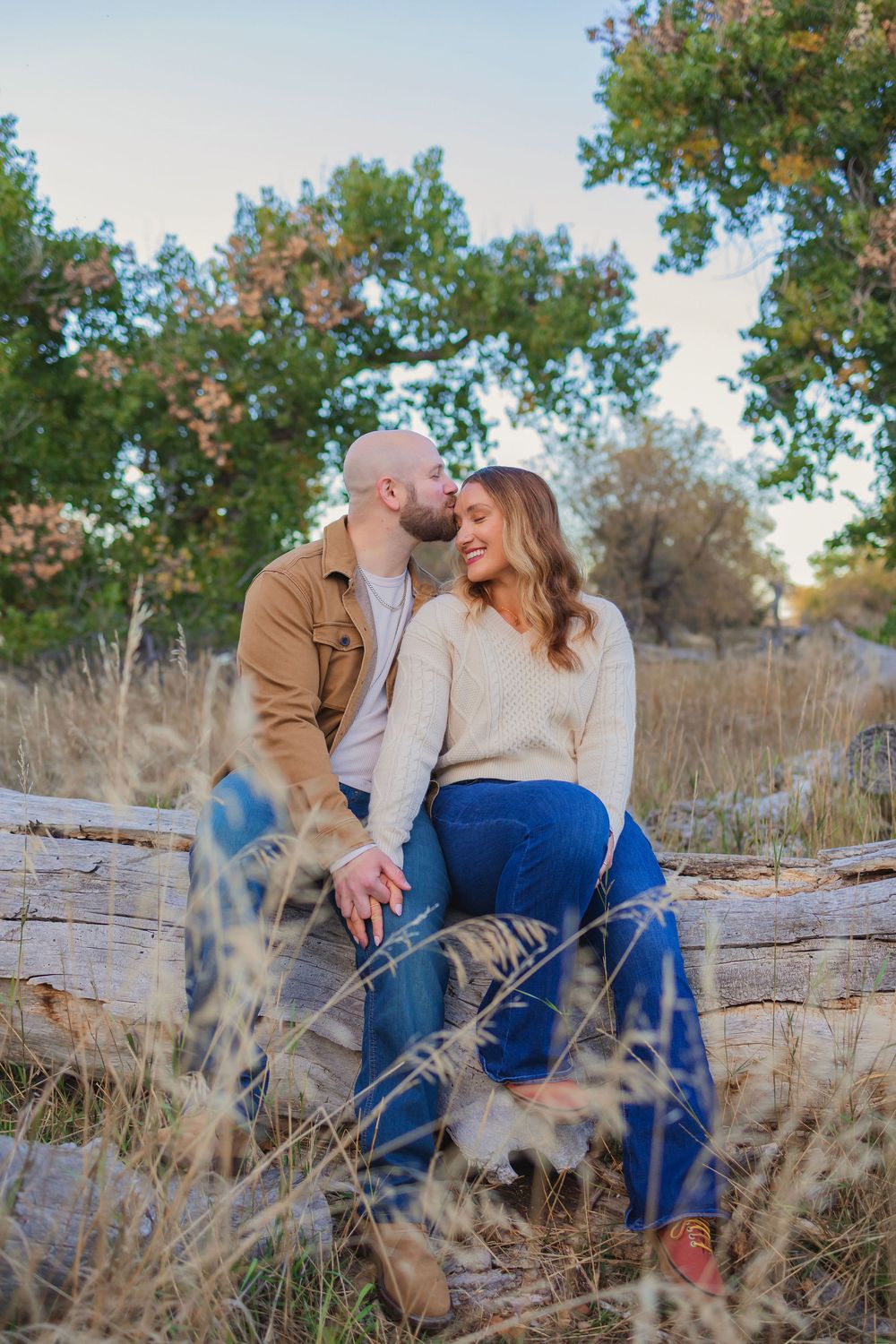 A romantic couple shares tender moments while sitting on a fallen tree trunk surrounded by autumn grasses and trees.