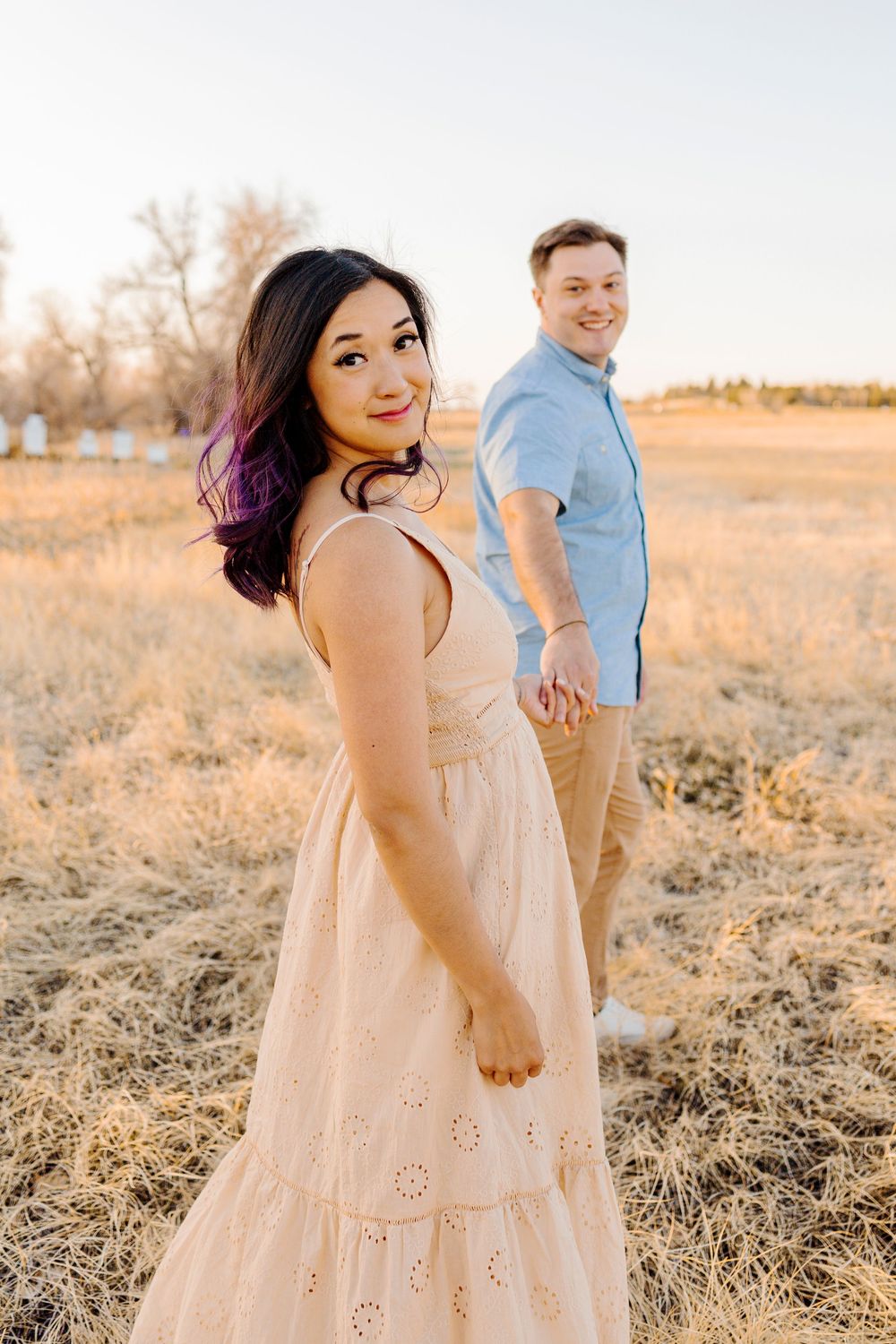 A couple poses together in a golden field at sunset wearing light colored clothing.