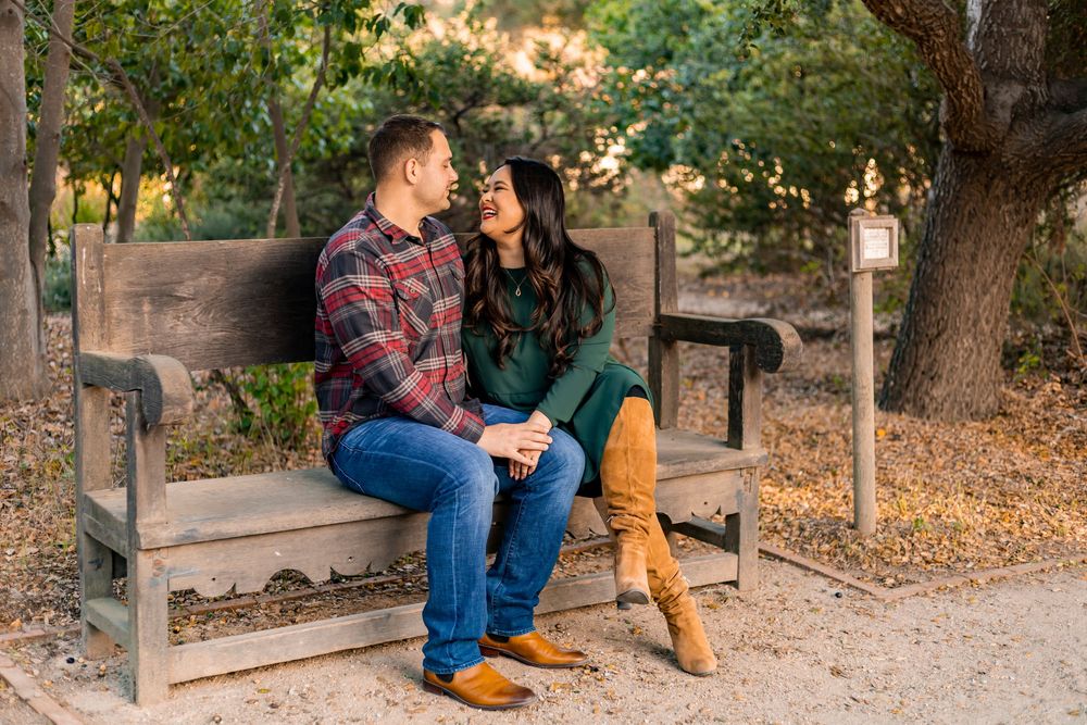 Two people sit on a wooden bench in a park setting during autumn with fallen leaves on the ground.