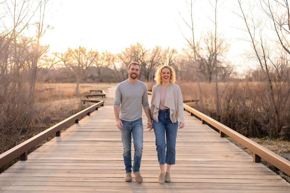 A couple walks hand in hand along a wooden boardwalk path at sunset through a natural landscape.