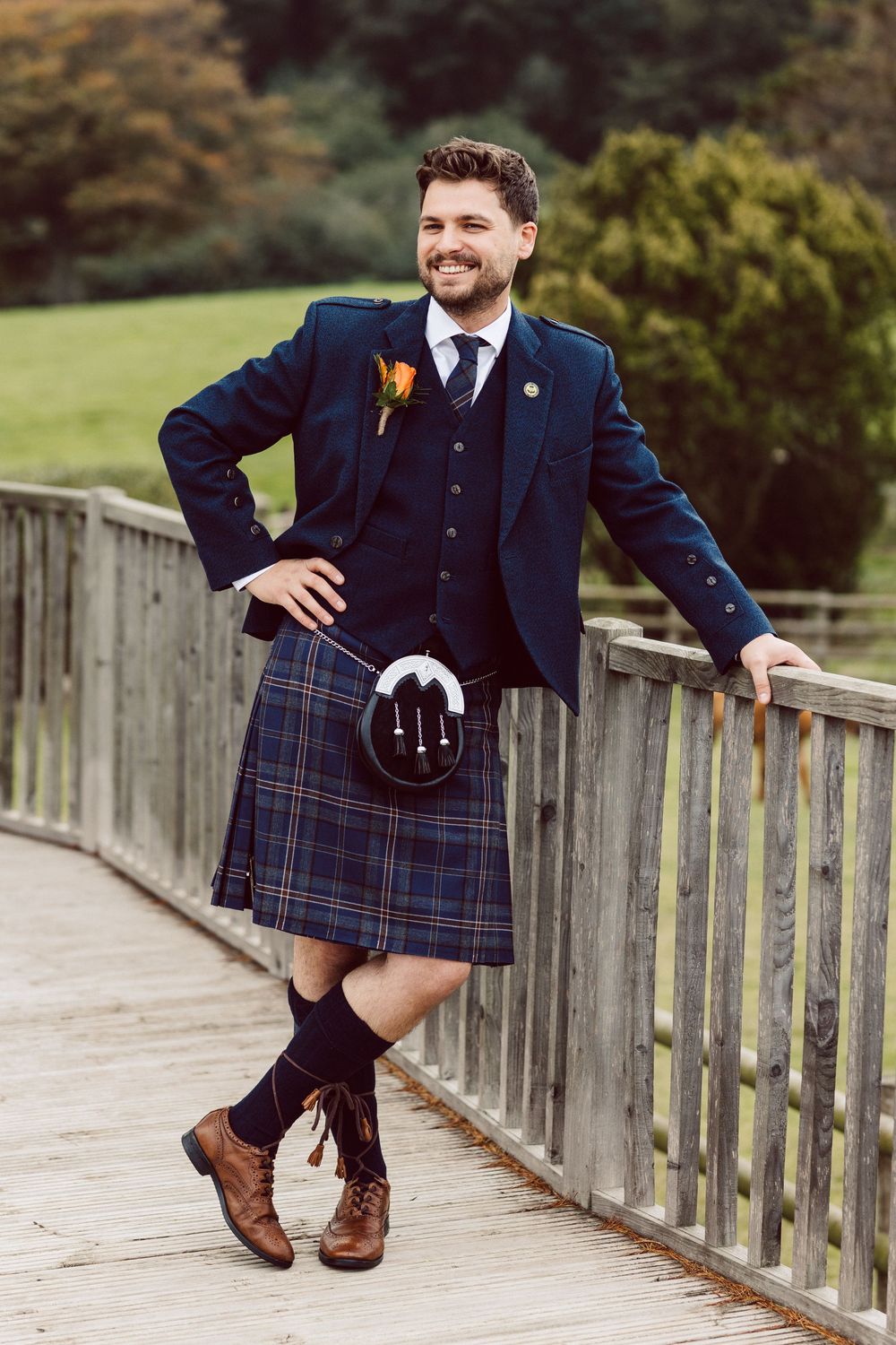 Men in traditional Scottish kilts and navy jackets posing playfully on a wooden bridge.