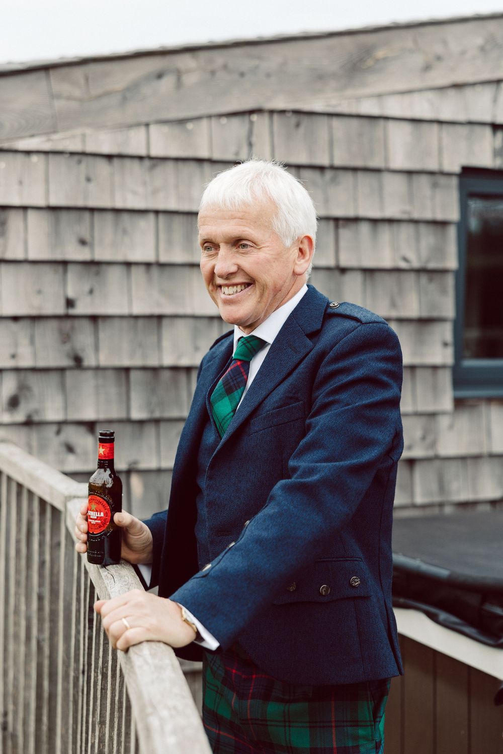 A person in a navy suit and green plaid vest smiles while holding a bottle outdoors.