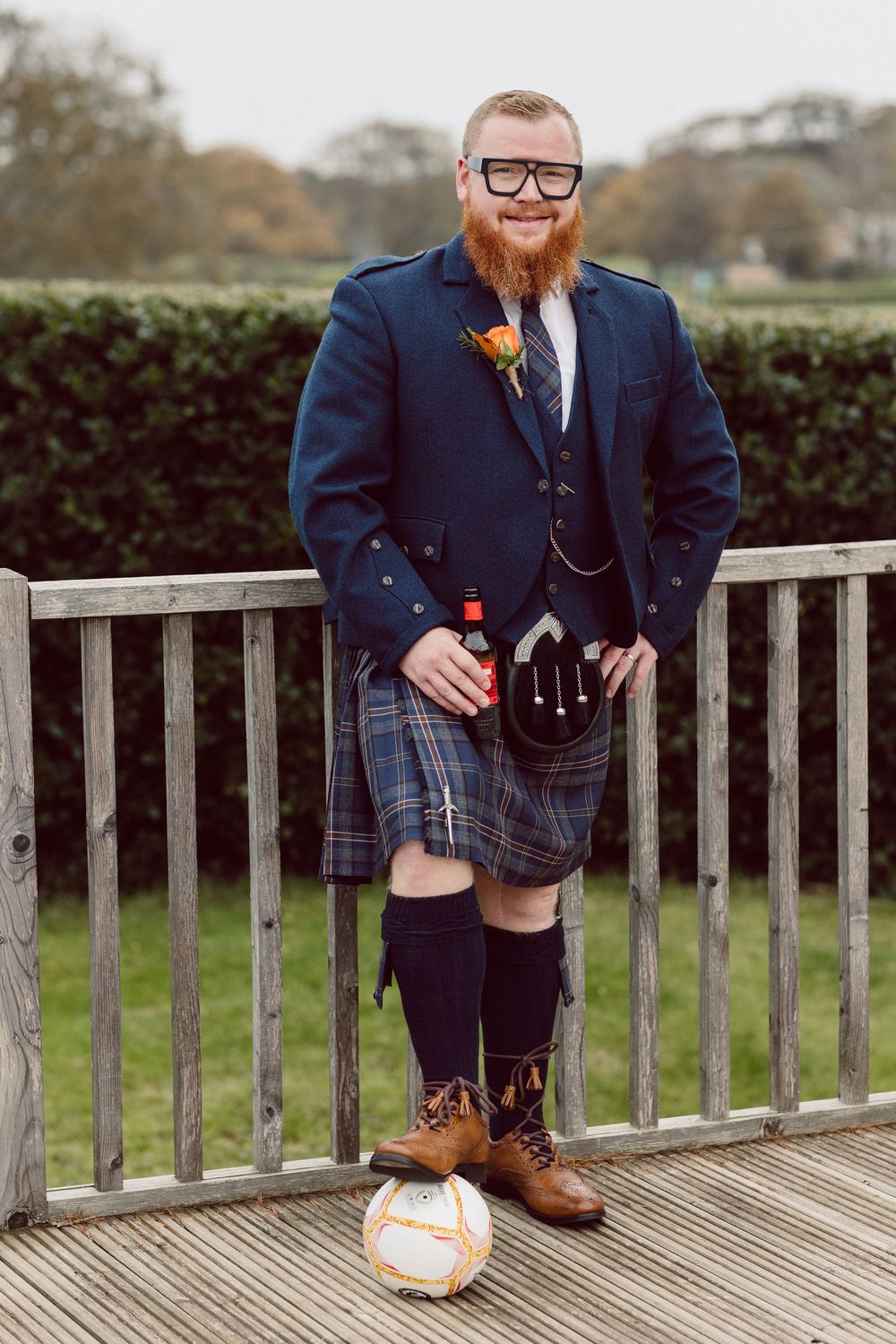 Person in traditional Scottish attire and glasses posing on wooden deck railing.