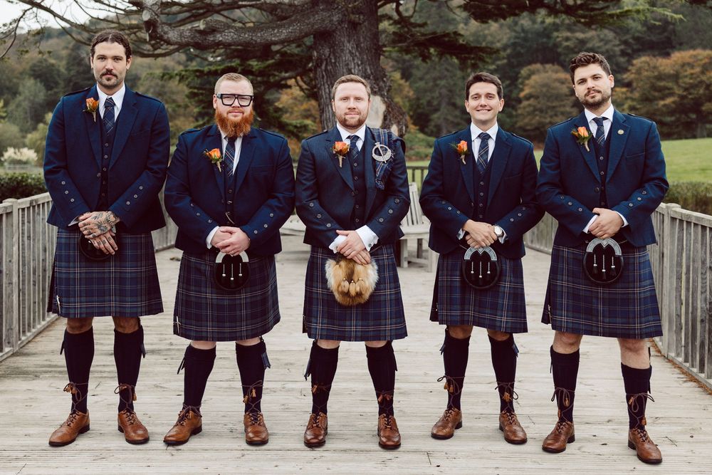 Five men in traditional Scottish kilts and formal attire stand together on a bridge in a garden setting.