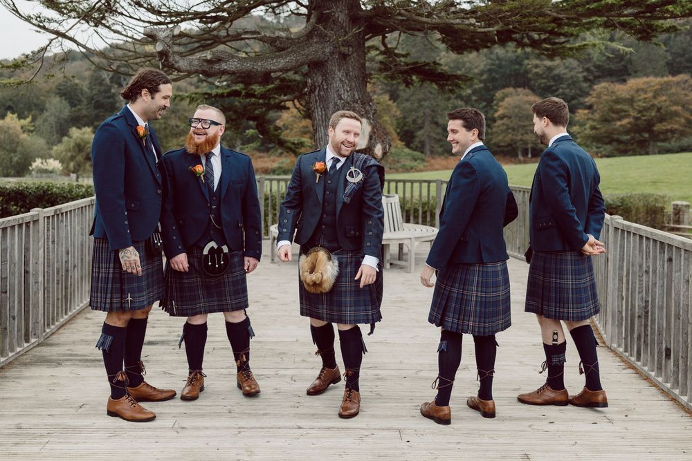 A group in matching blue plaid suits and brown shoes stand together on a wooden bridge in a natural setting.
