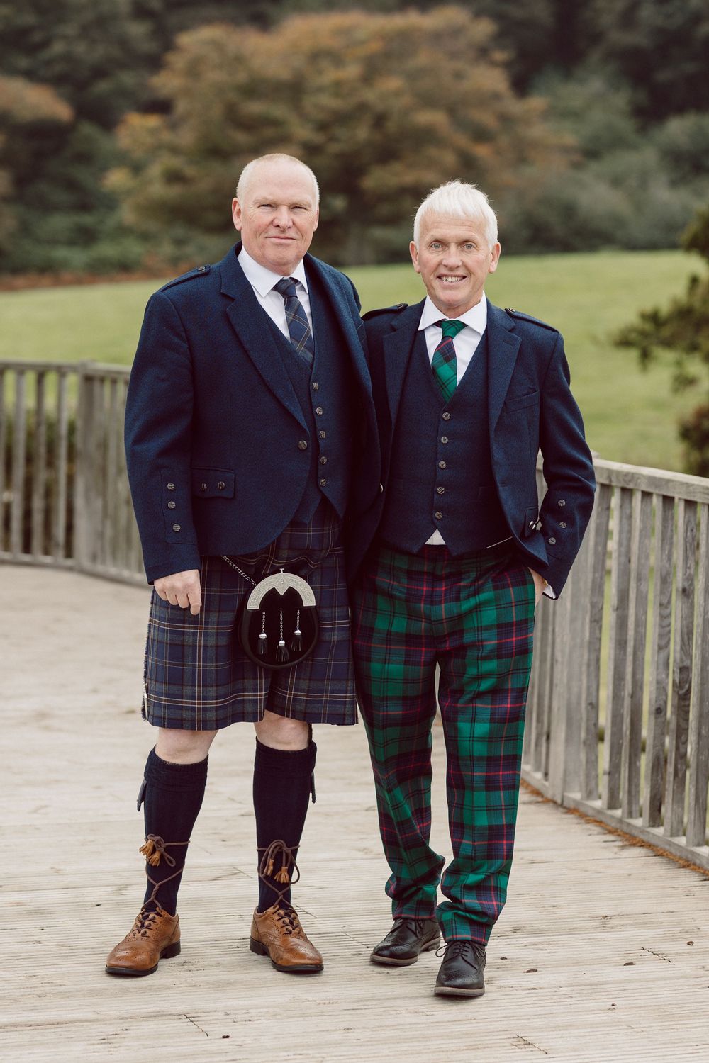 Two people in traditional Scottish kilts and formal jackets stand together on a wooden bridge.