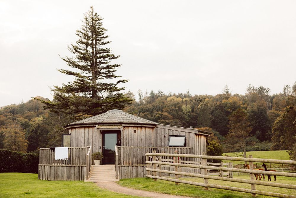 Panoramic view of wooden outbuildings and fencing nestled among evergreen trees on rural property.