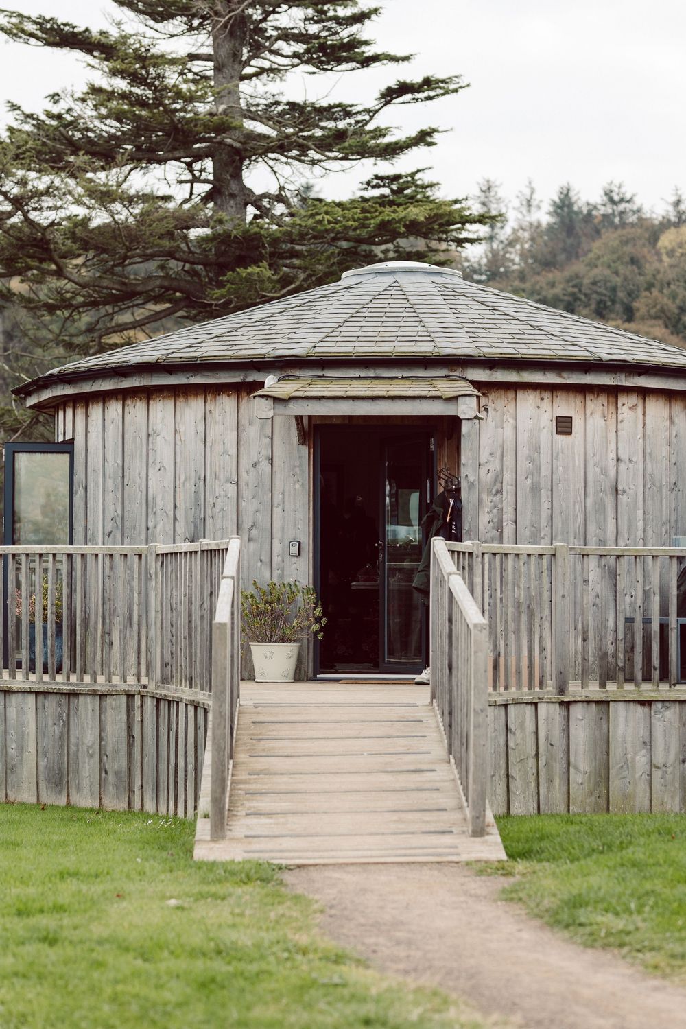 Rustic wooden cabin with ramp entrance and weathered siding surrounded by evergreen trees.