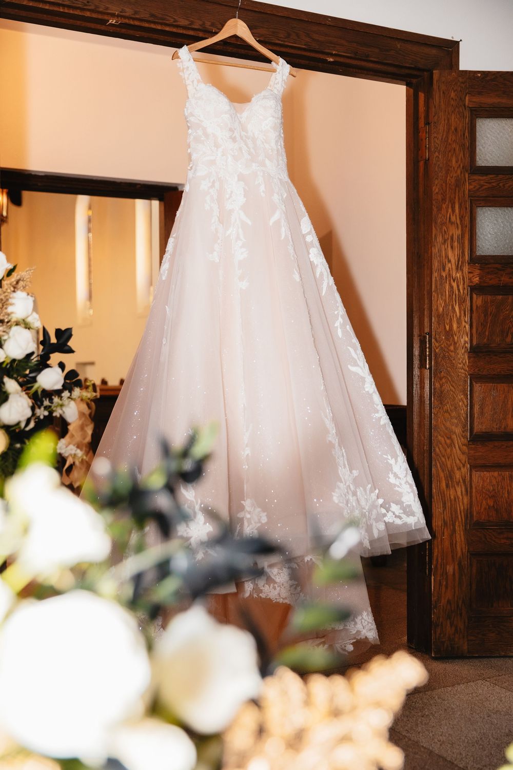 A delicate white wedding dress with lace appliques hangs on a wooden hanger against a warm-toned wall with floral decor.