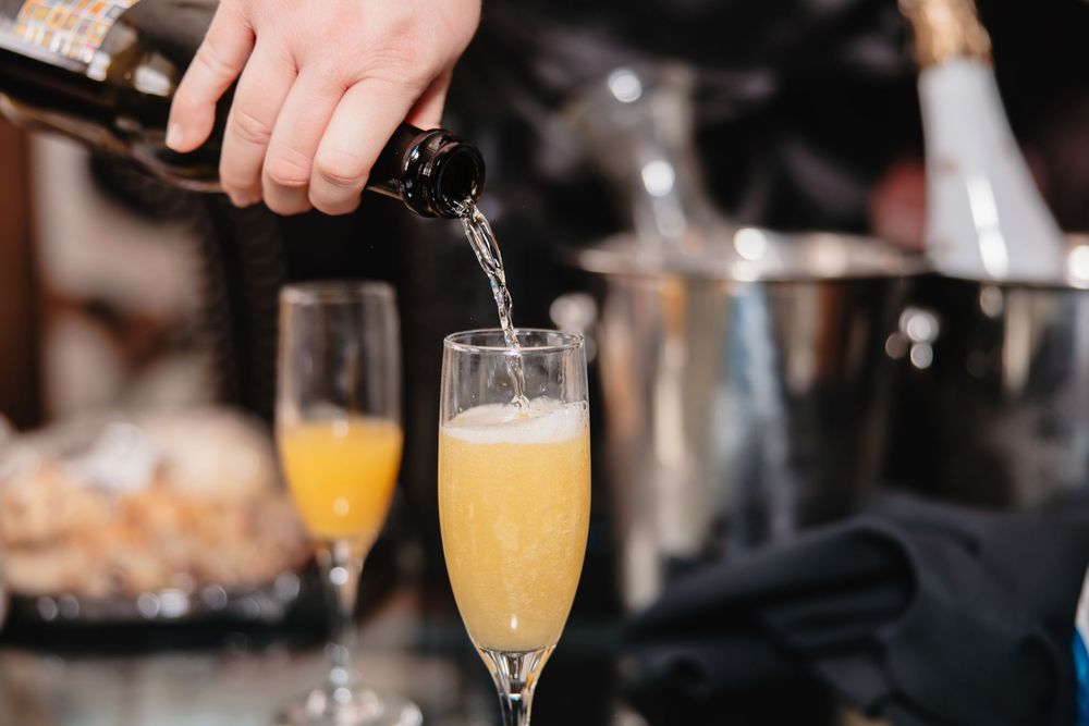 Champagne being poured into a mimosa cocktail glass with orange juice at a bar service station.