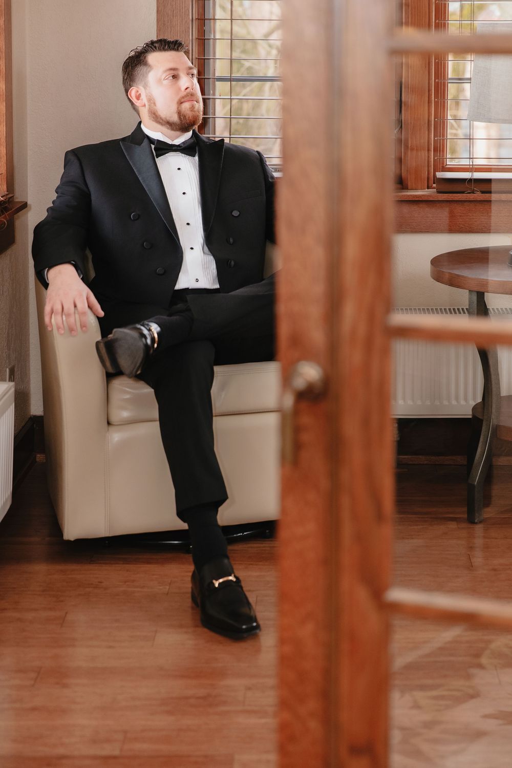 Person in formal black tuxedo lounges on elegant wooden furniture in an interior setting.