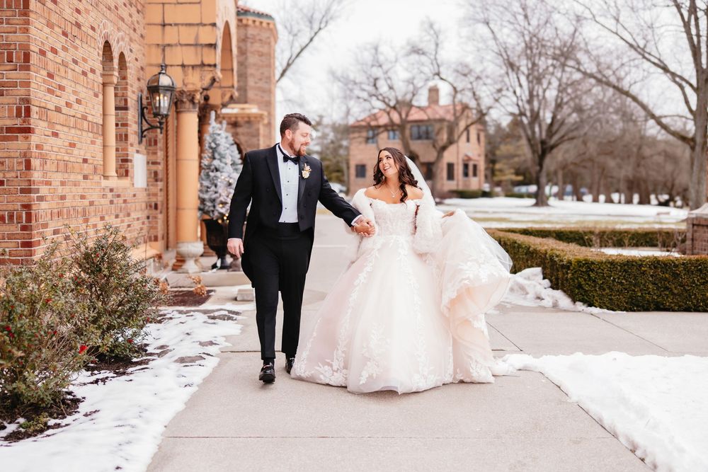 Couple walks together on snowy path outside brick building during winter wedding.