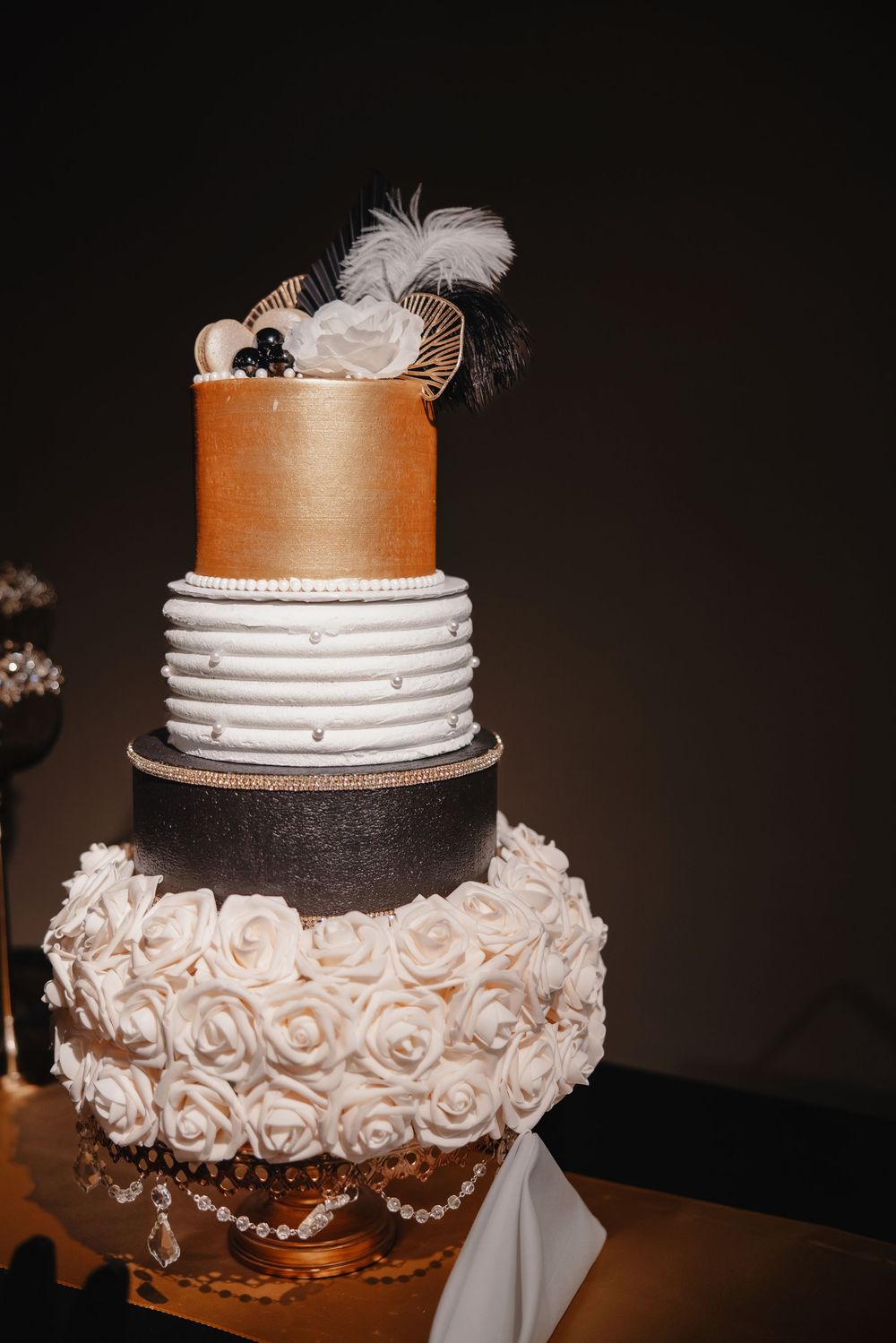 A three-tiered wedding cake decorated with white roses, orange fondant, and delicate pearl details on a dark background.