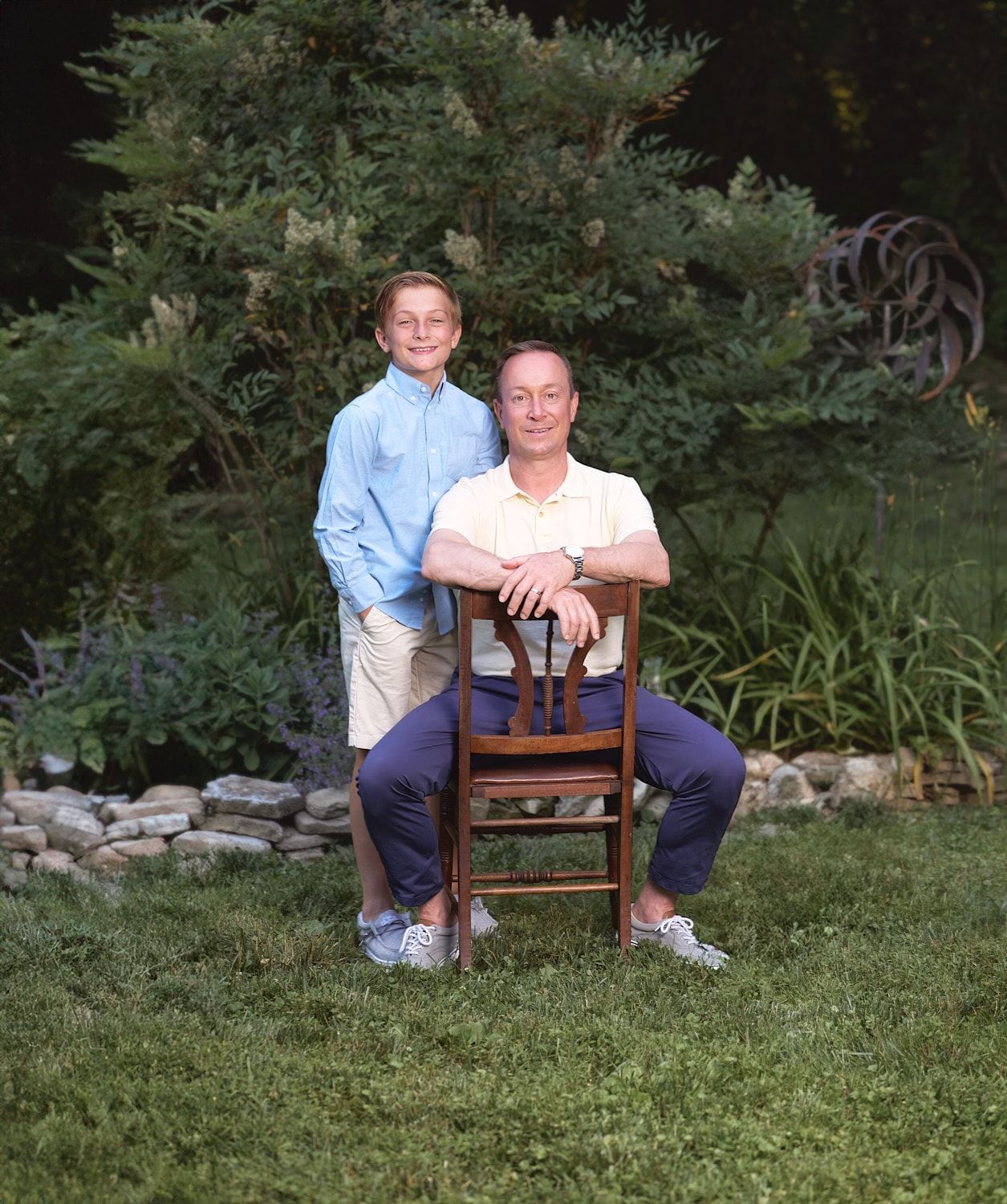 Dad straddling an antique dining chair with tween son standing aside him in a back yard scene