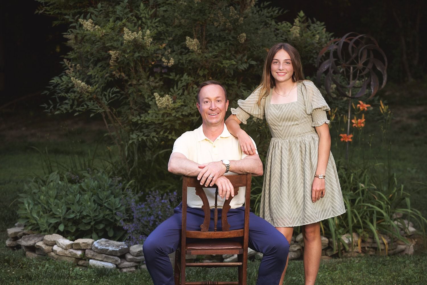 Dad sitting cowboy style on East Lake style chair with teen daughter standing and leaning on his shoulder with a happy smile.