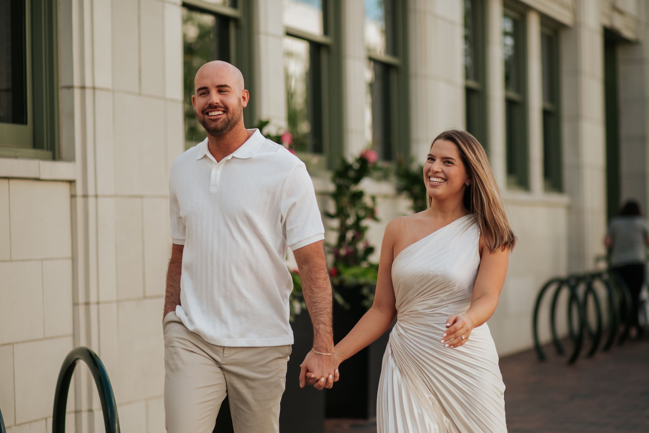 Couple in white outfits holding hands and walking together along a building exterior.