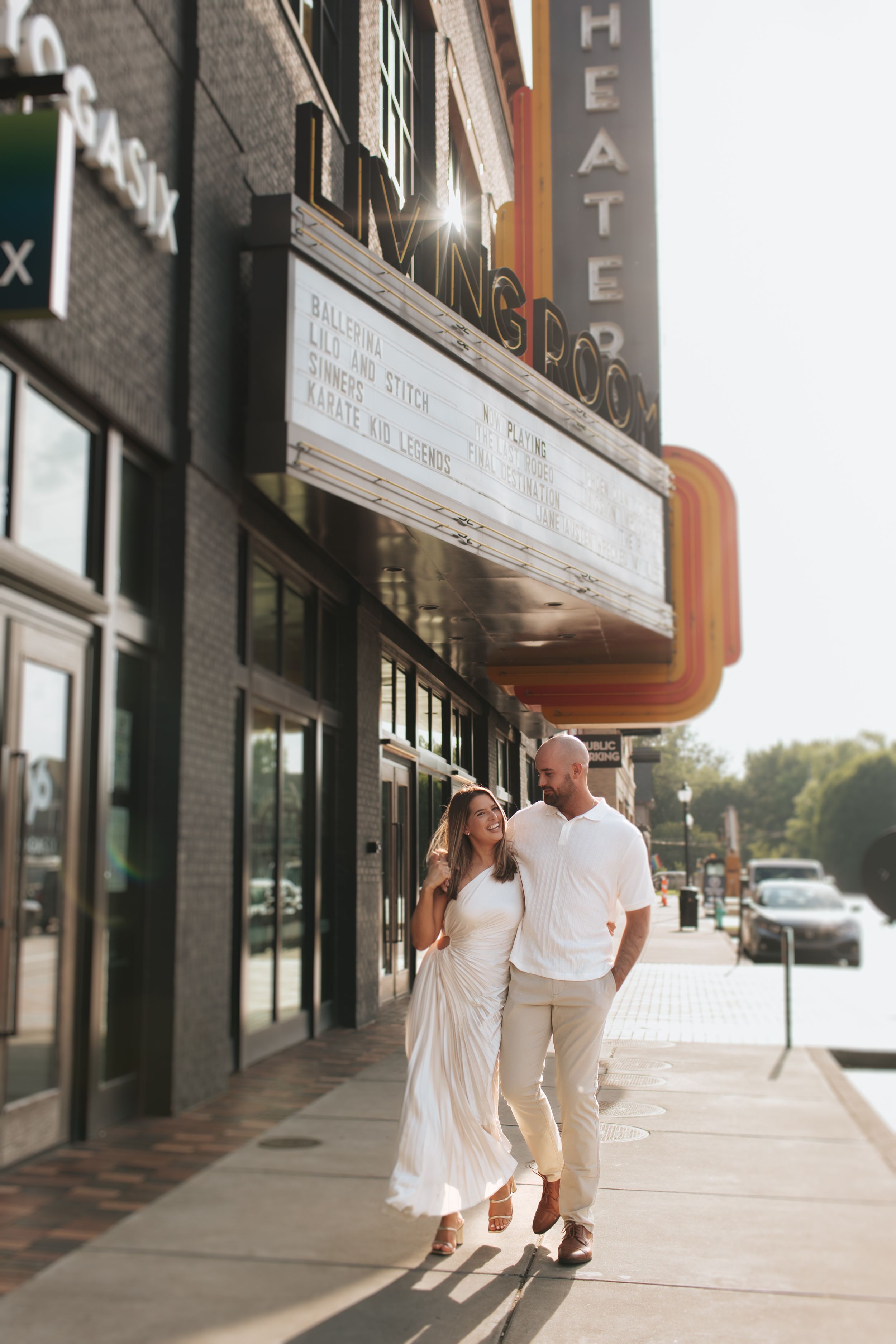 A couple in white outfits walks together along a storefront sidewalk in warm evening light.