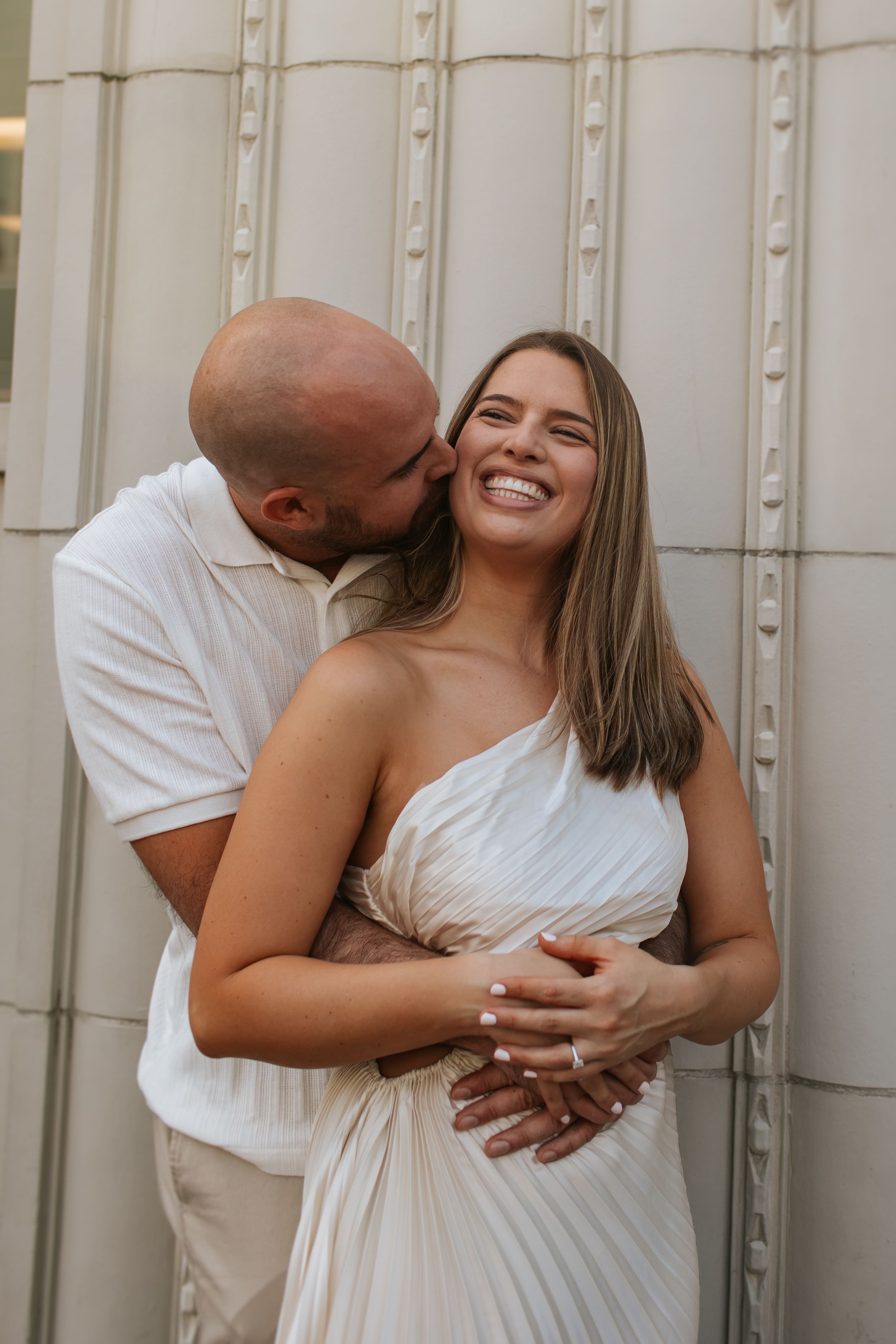 A romantic couple in white clothing embraces against a light colored architectural wall.