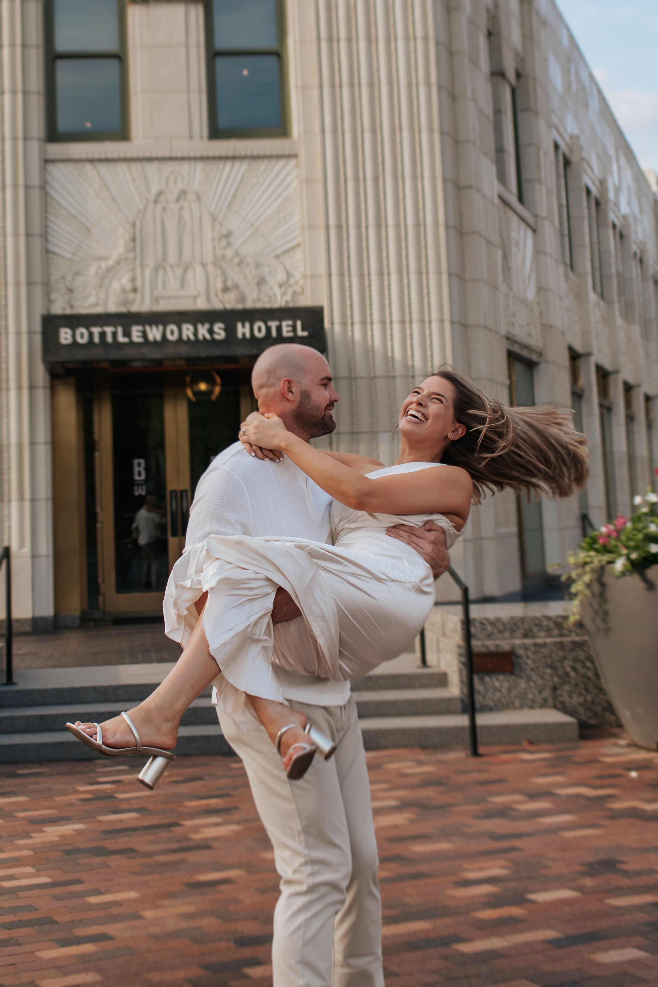 A couple in white outfits dancing and embracing outside the Bottleworks Hotel building.
